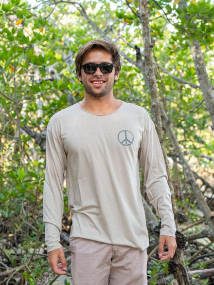 Man standing among tree branches wearing a tan long-sleeve shirt with a peace symbol and light brown shorts.