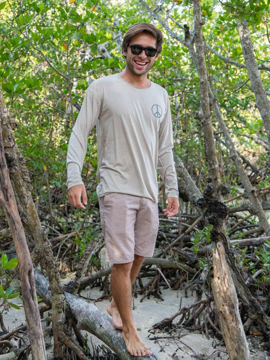 Man standing among tree branches wearing a tan long-sleeve shirt with a peace symbol and light brown shorts.