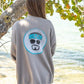 Woman wearing a gray sweatshirt with a logo, standing by a tree on a beach.
