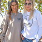 Two women wearing sweatshirts with text, standing by a tree on a beach.