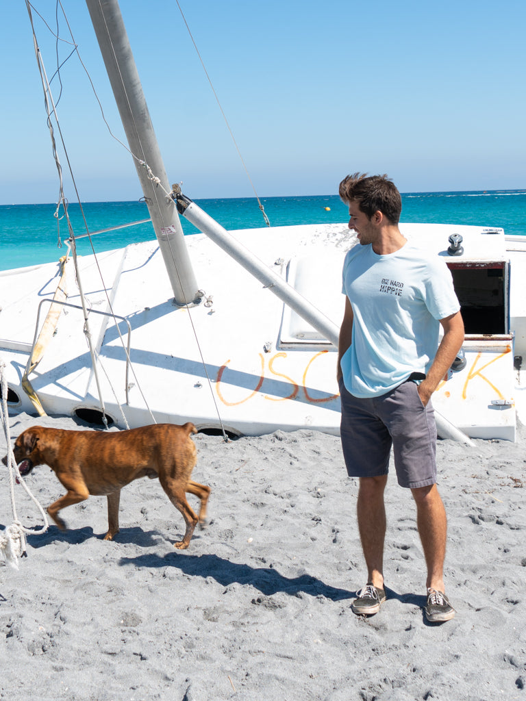 Man standing by a boat wearing a light blue t-shirt with a logo, looking at the ocean.