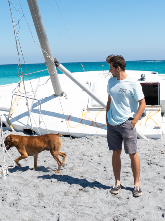 Man standing by a boat wearing a light blue t-shirt with a logo, looking at the ocean.