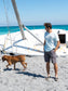 Man standing by a boat wearing a light blue t-shirt with a logo, looking at the ocean.