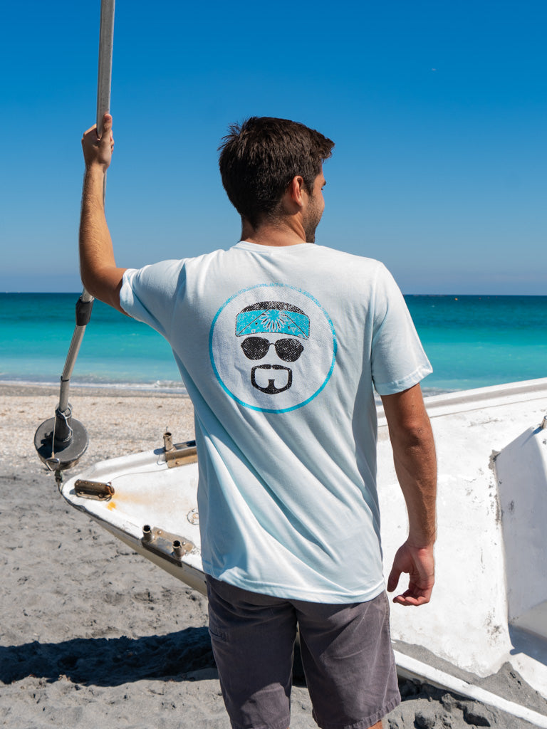 Man standing on a boat wearing a light blue t-shirt with a logo, looking at the ocean.