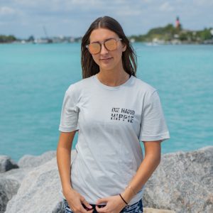 Woman wearing a gray t-shirt with text and graphics, standing on rocks by a body of water.