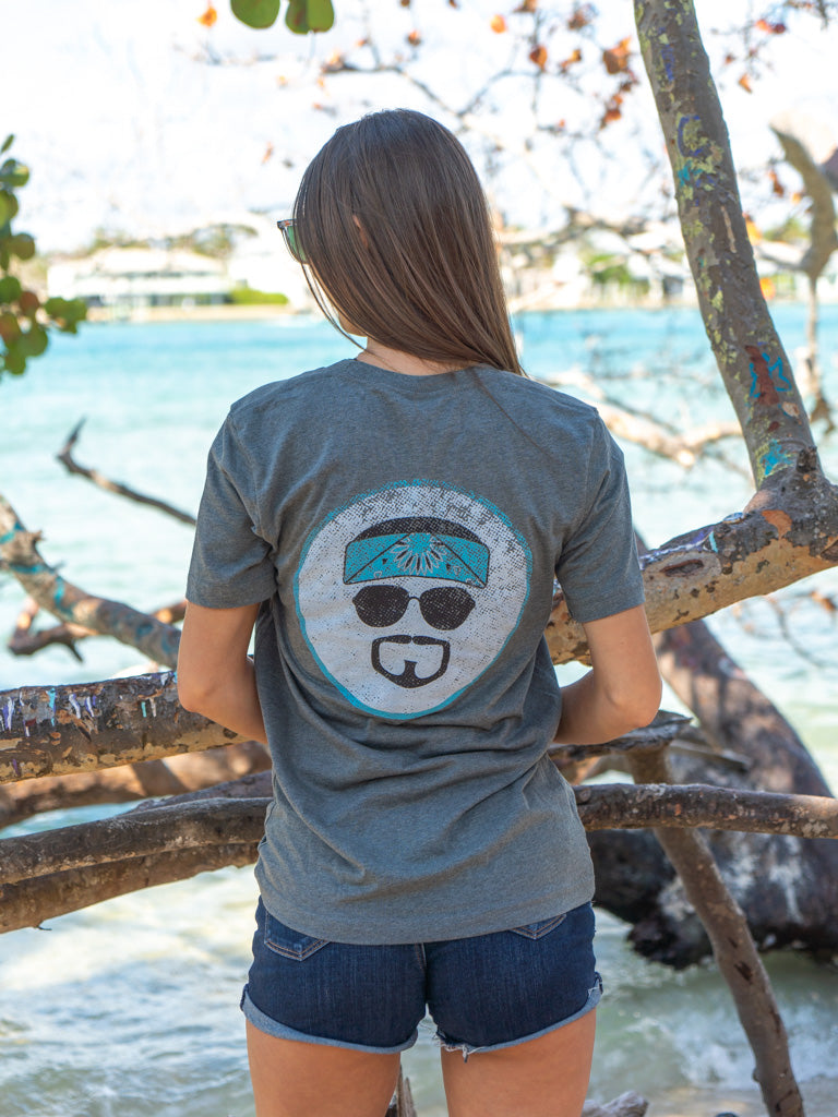 Woman wearing a gray t-shirt with a logo featuring sunglasses and a bandana, standing by rocks and water.