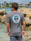 Man wearing a gray t-shirt with a circular design featuring sunglasses and a face, standing by rocks and water.