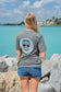 Woman wearing a gray t-shirt with a logo, standing by rocks and water.