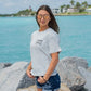 Woman wearing a gray t-shirt with text and graphics, standing on rocks by a body of water.