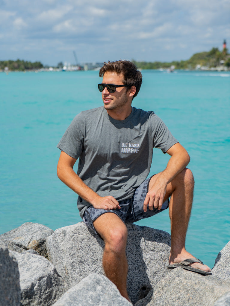 Man wearing a gray t-shirt with the words 'Die Hard Hippie', sitting on rocks by water.