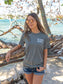 Woman wearing a gray t-shirt with the words 'Die Hard Hippie', standing by rocks and water.