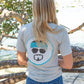 Woman wearing a gray t-shirt with text and graphics, standing on rocks by a body of water.
