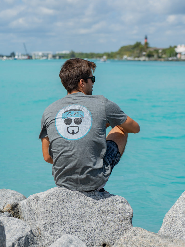 Man wearing a gray t-shirt with a circular design featuring sunglasses and a face, sitting on rocks by water.