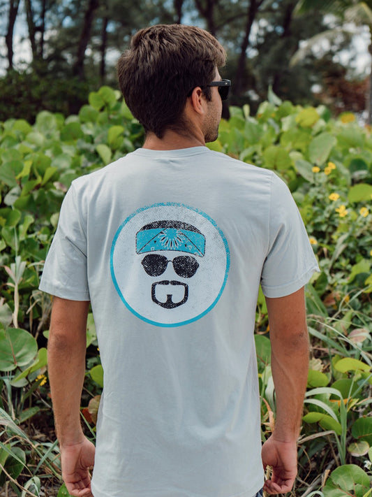Man wearing a gray t-shirt with graphic, standing by greenery in a beach setting