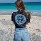 Woman wearing a black t-shirt with text and graphics, standing on rocks by a body of water.