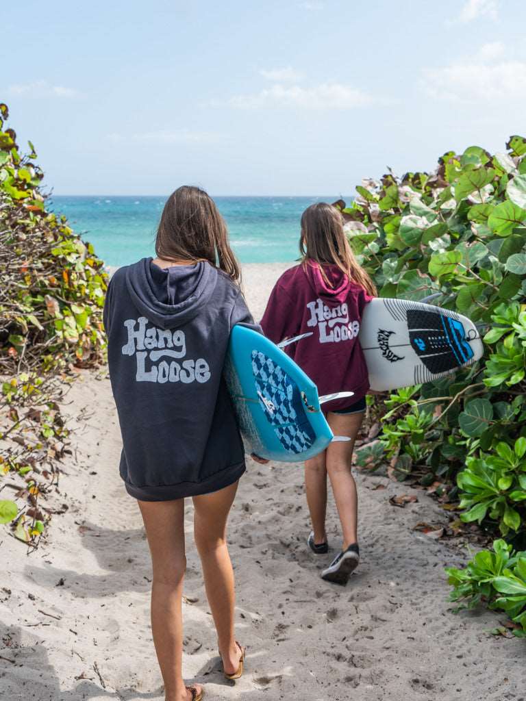 Two people walking on a beach with surfboards, wearing 'Hang Loose' hoodies.