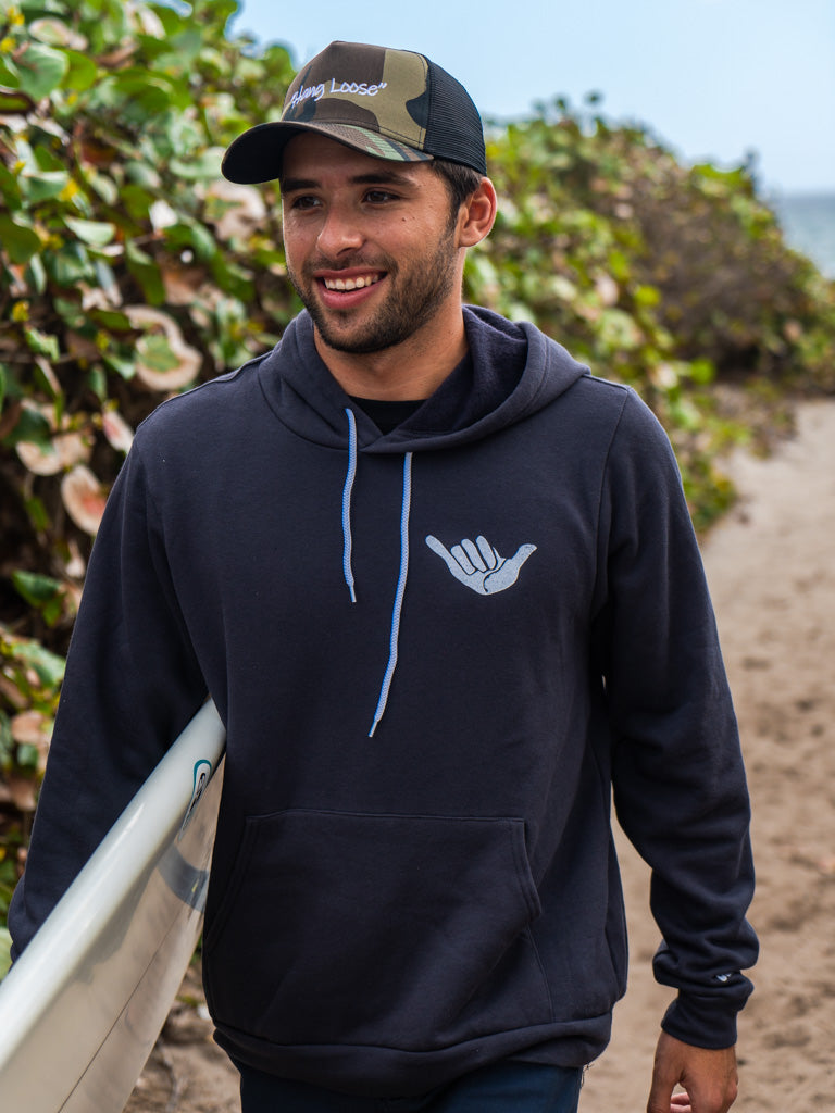 Man walking on beach wearing dark grey hoodie with a hang loose sign on the front