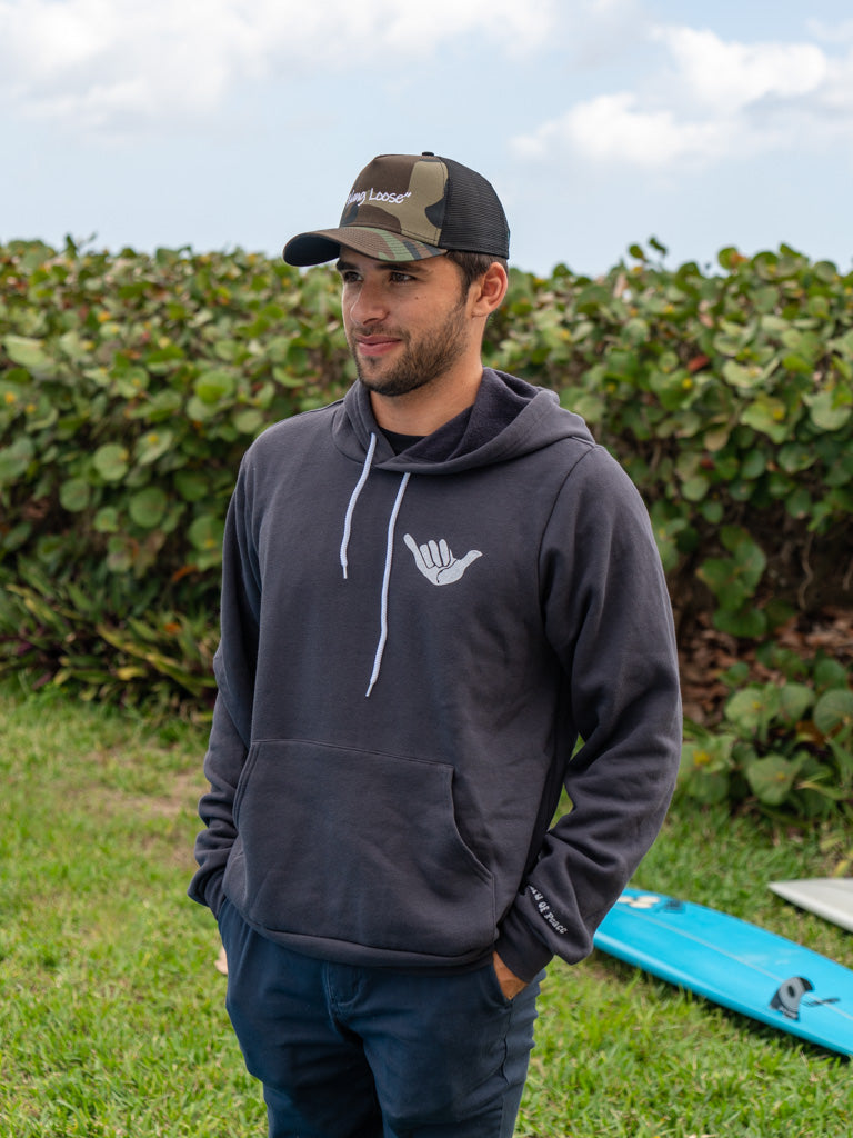 Man walking on beach wearing dark grey hoodie with a hang loose sign on the front with greenery in the background