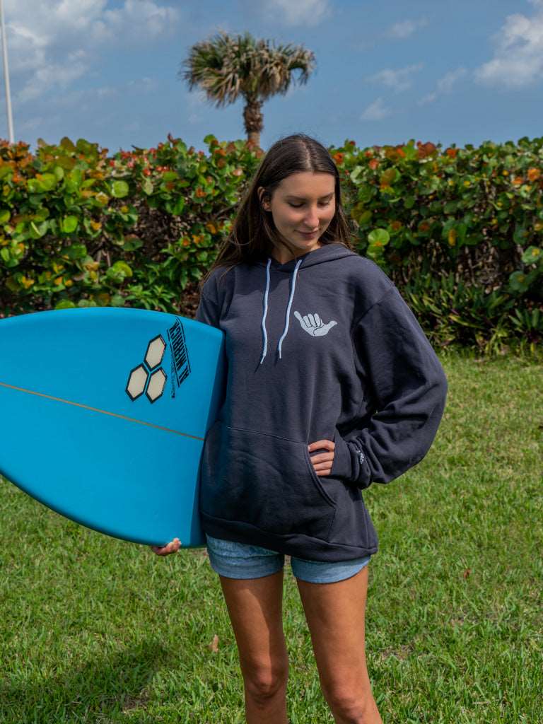 Woman holding a surfboard wearing dark grey hoodie with a hang loose sign on the front with greenery in the background
