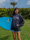 Woman holding a surfboard wearing dark grey hoodie with a hang loose sign on the front with greenery in the background