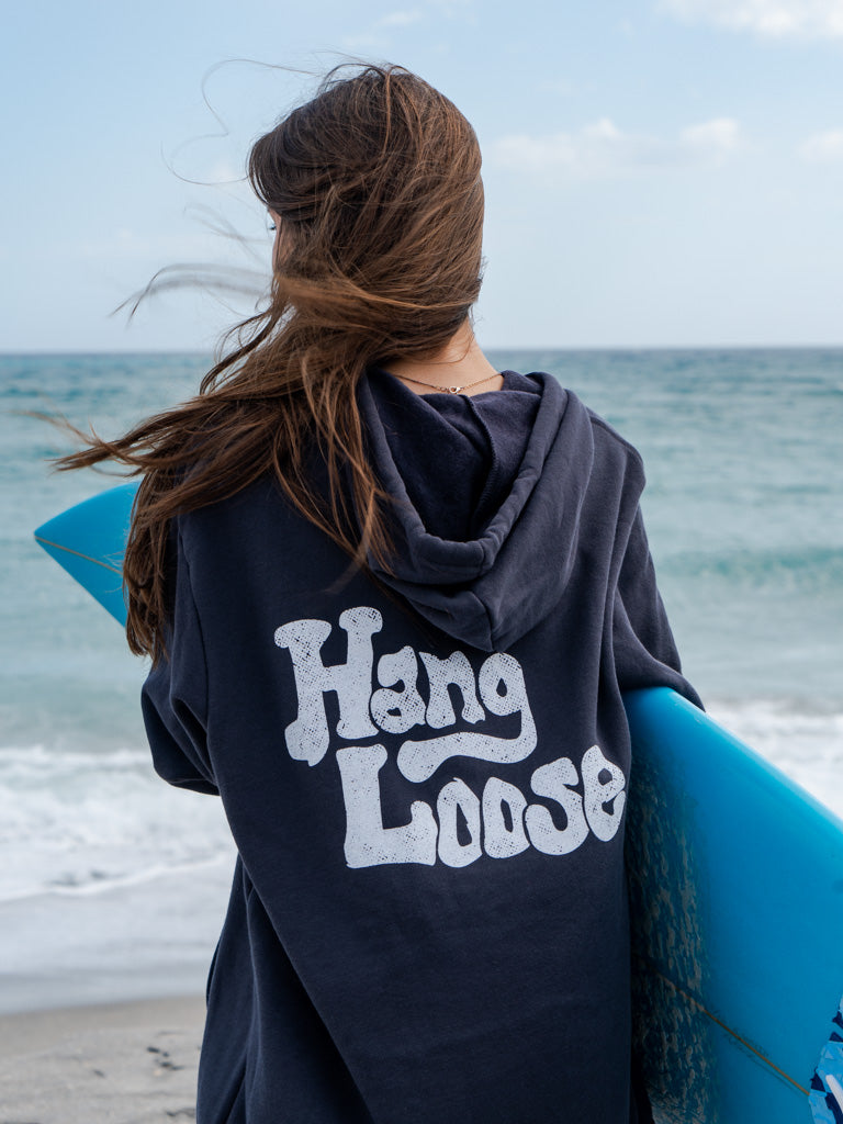 Woman holding a surfboard wearing dark grey hoodie with the words 'Hang Loose' on the back with the ocean in the background