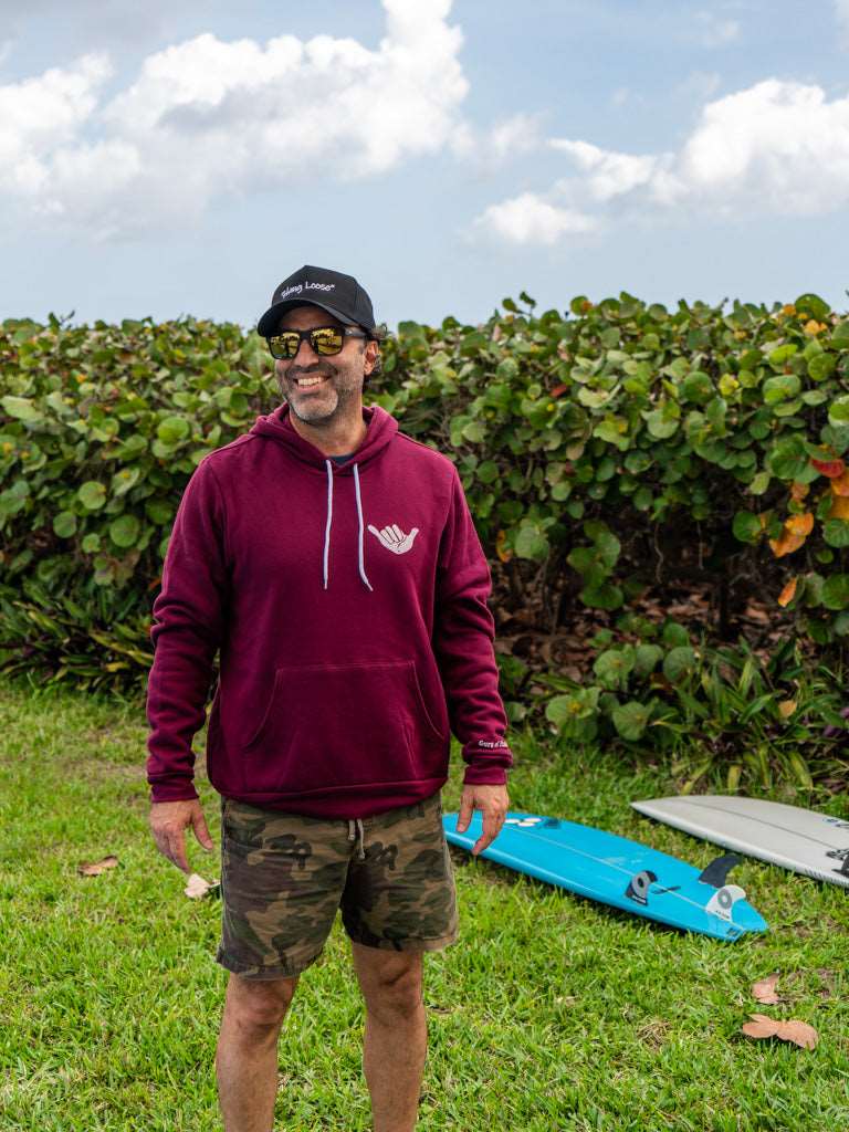 Man wearing maroon hoodie with a hang loose sign on the front, standing with greenery in the background