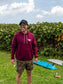 Man wearing maroon hoodie with a hang loose sign on the front, standing with greenery in the background