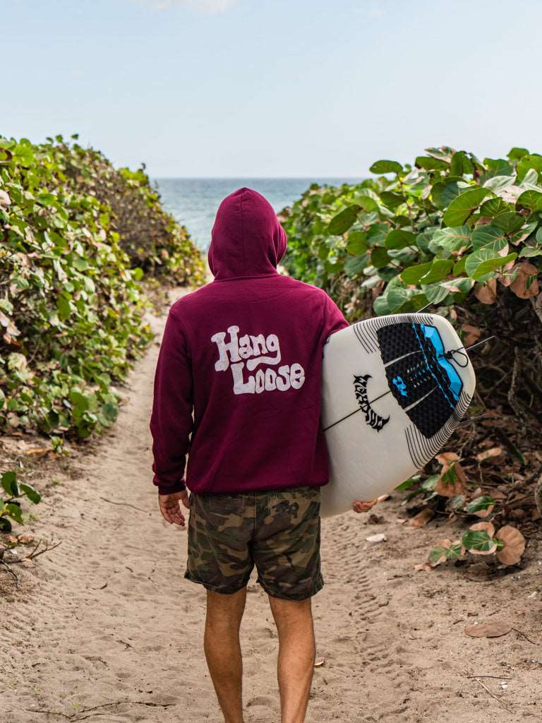Man holding a surfboard wearing maroon hoodie with the words 'Hang Loose' on the back with the ocean in the background
