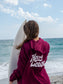 Woman holding a surfboard wearing maroon hoodie with the words 'Hang Loose' on the back with the ocean in the background