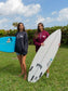Two women holding a surfboard wearing a maroon and dark grey hoodie with a hang loose sign on the front with greenery in the background