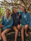 Three women wearing grey and blue hoodies with a logo, sitting together outdoors.