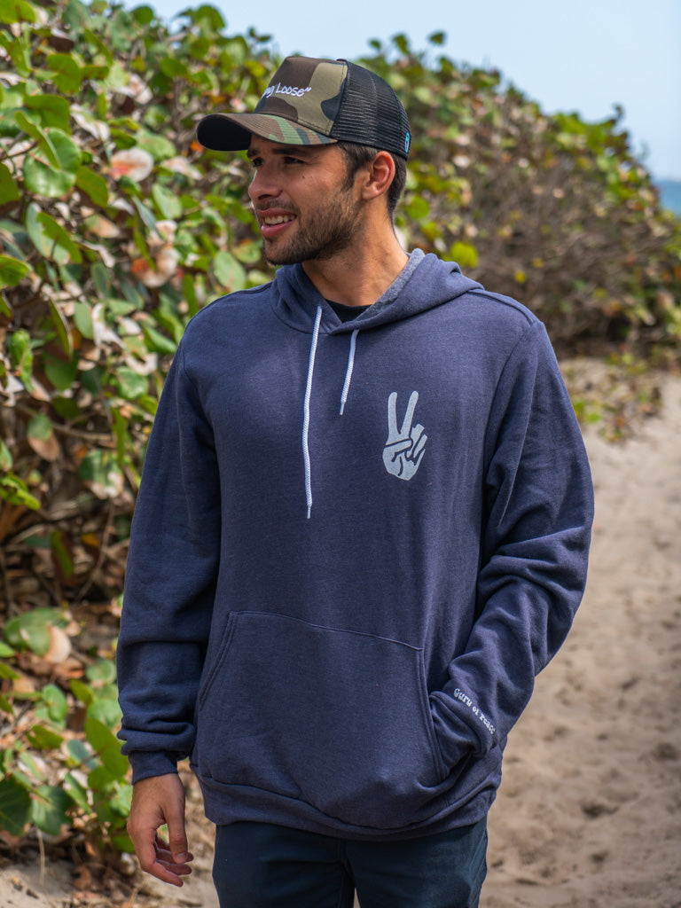 Man wearing navy hoodie with a hand signing peace, walking on a beach