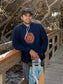 Man wearing a hoodie with a sun design on it, holding a skateboard on a wooden deck with trees in the background