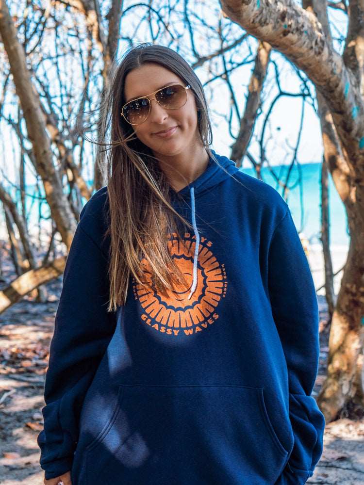 Woman wearing navy hoodie with sun design, on a beach with trees in the background