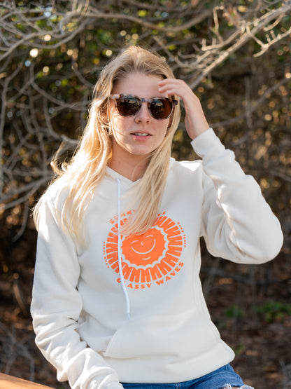 Woman wearing white hoodie with sun design, sitting on a wooden bench with trees in the background