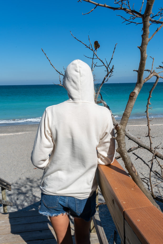Woman wearing white hoodie, standing on a wooden deck with a beach in the background