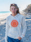 Woman wearing white hoodie with sun design, standing on a beach with trees in the background