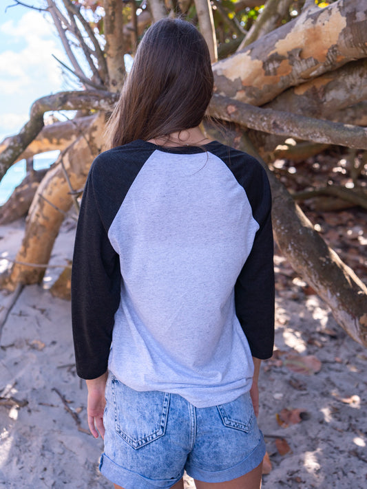 Woman wearing a black and white baseball shirt, standing by branches on a beach.