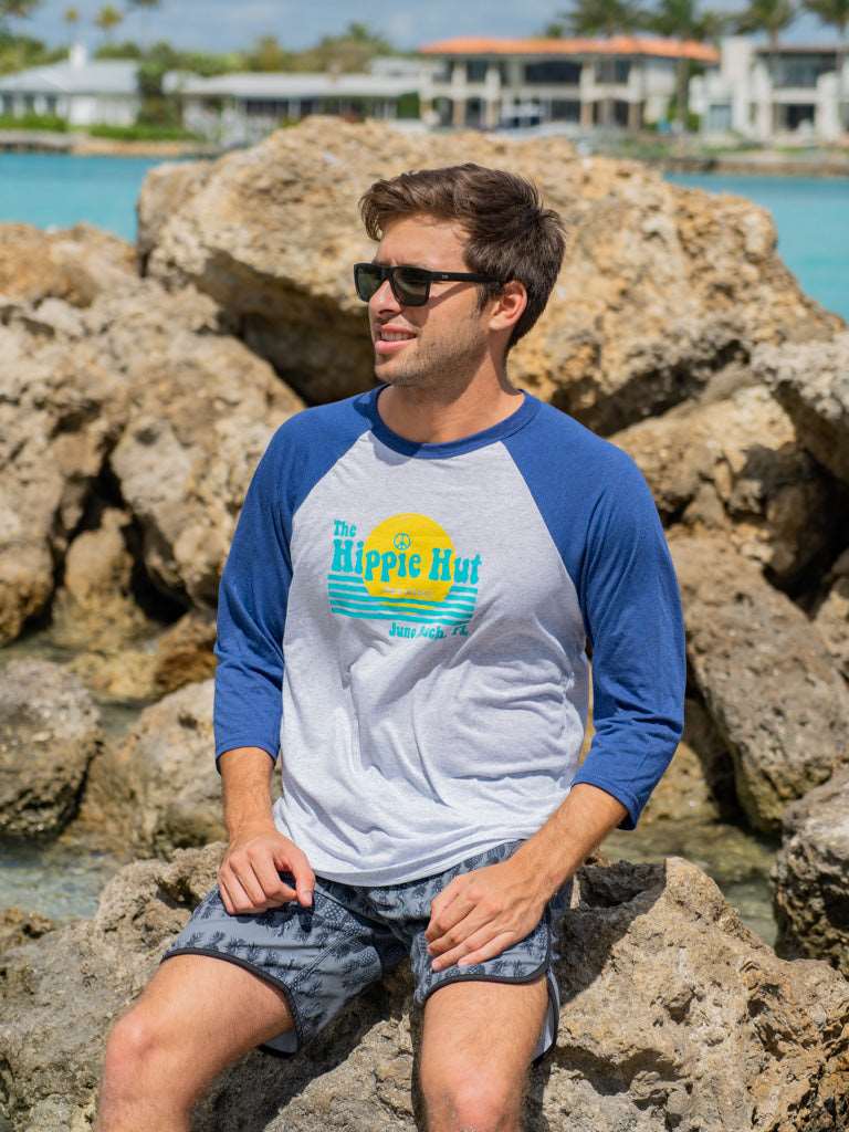 Man wearing a blue and white baseball shirt with a logo, sitting on rocks by a waterfront.