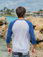 Man wearing a blue and white baseball shirt, standing by rocks by a waterfront.