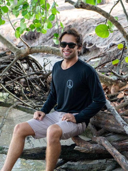 Man sitting among tree branches wearing a black long-sleeve shirt with a peace symbol and light gray shorts.