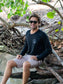 Man sitting among tree branches wearing a black long-sleeve shirt with a peace symbol and light gray shorts.