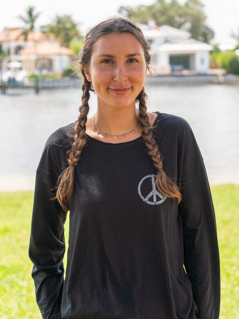 Woman standing by a body of water wearing a black long-sleeve shirt with a peace symbol.