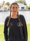 Woman standing by a body of water wearing a black long-sleeve shirt with a peace symbol.