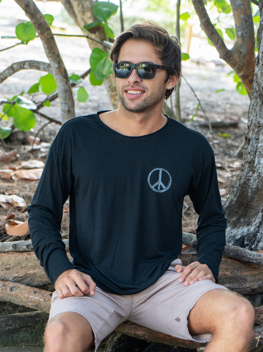Man sitting among tree branches wearing a black long-sleeve shirt with a peace symbol and light gray shorts.