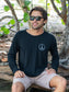 Man sitting among tree branches wearing a black long-sleeve shirt with a peace symbol and light gray shorts.