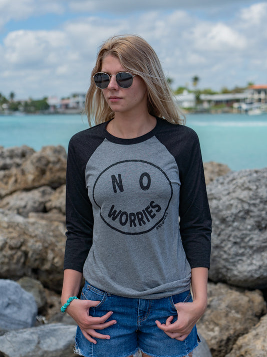 Woman wearing a 'No Worries' shirt standing in front of rocks and the ocean.