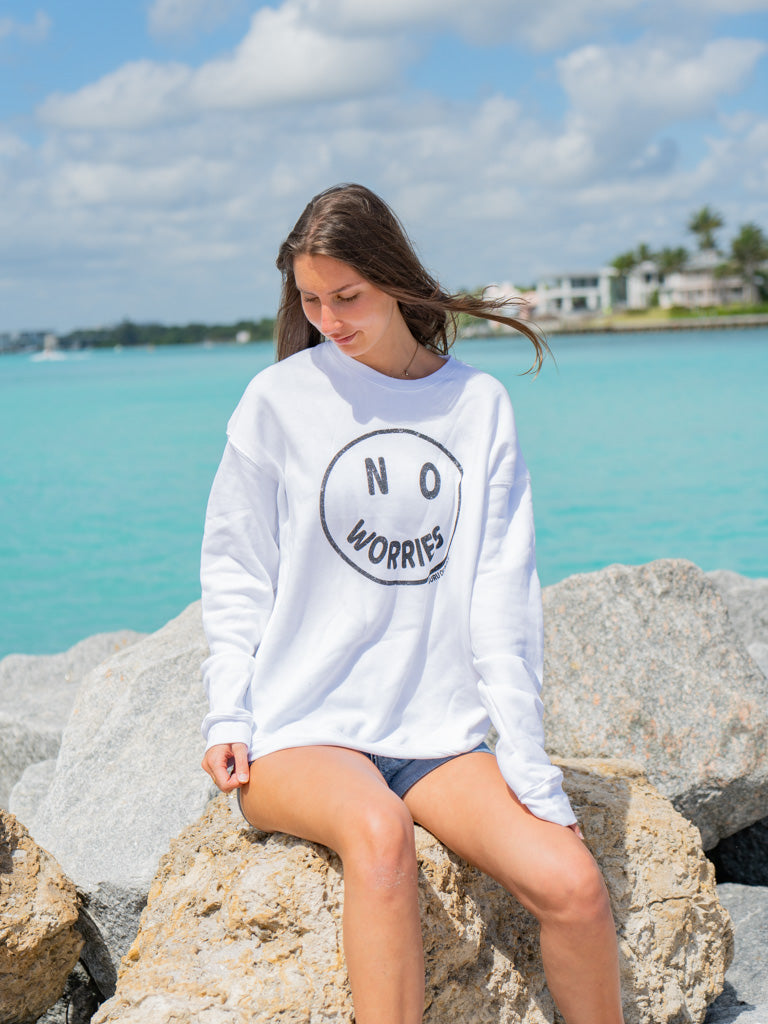 Woman wearing a white sweatshirt with the words 'No Worries' by a body of water.