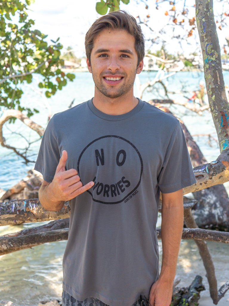 Man wearing a gray t-shirt with 'No Worries' graphic, standing by a body of water.