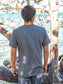 Man wearing a gray t-shirt with 'No Worries' graphic, standing by a body of water.